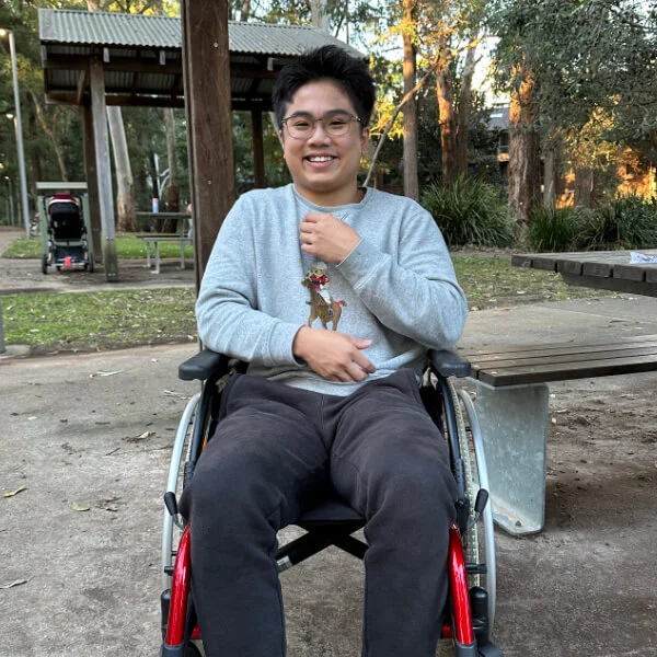 Young man in a wheelchair smiling at the camera in an outdoor setting.