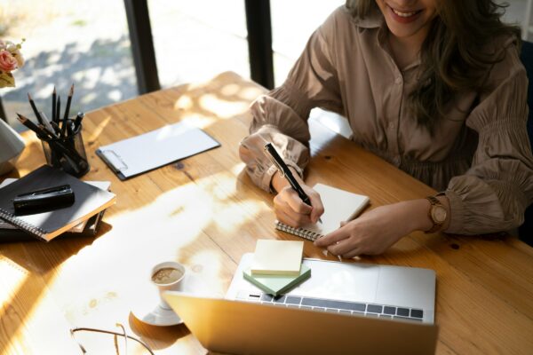 Woman smiling while taking notes at a desk with a laptop and coffee, finding the best support through NDIS Plan Management.
